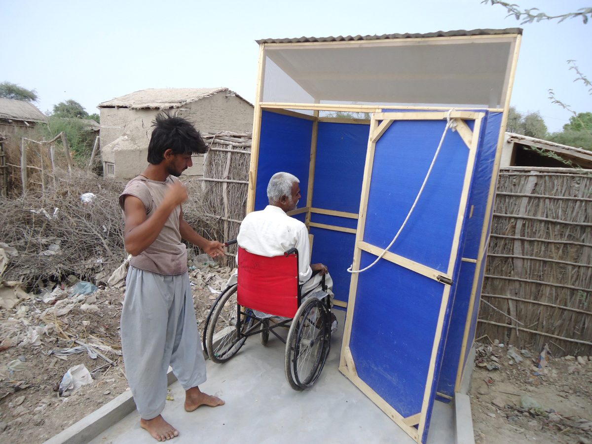A man in a wheelchair is entering a temporary latrine. The door has a rope hanging from the top for make easy the closing