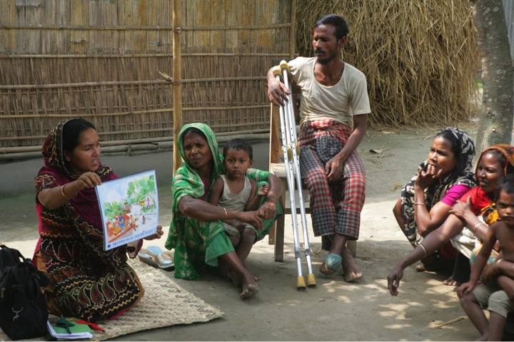 মাটিতে বসা একজন নারী কর্মী ফ্লিপচার্টের ছবি দেখিয়ে তারই মতো মাটিতে বসা একদল নারীকে তথ্য দিচ্ছেন এবং ক্রাচ ব্যবহারকারী একজন পুরুষ কাছেই একটি চেয়ারে বসে তাদের সাথে আলোচনায় অংশ নিচ্ছেন। নারীদের কোলে তাদের সন্তানেরা রয়েছে।