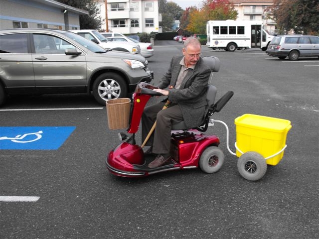 Seorang laki-laki pengguna kursi roda elektrik dengan gerobak terpasang di belakangnya. Gerobak berisi sebuah kotak.