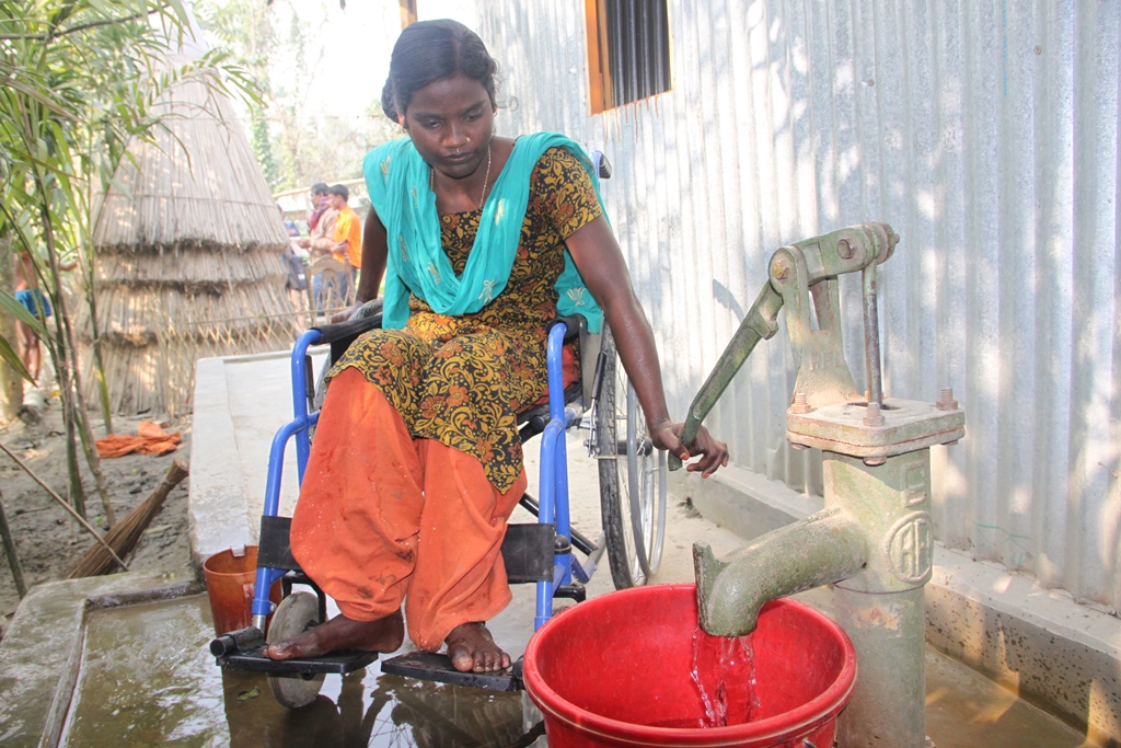 Wanita di kursi roda di Bangladesh menggunakan pompa tangan untuk mendapatkan air.