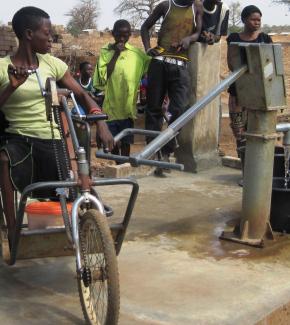 Seorang perempuan menggunakan sepeda roda tiga untuk mengoperasikan gagang pompa yang diperpanjang