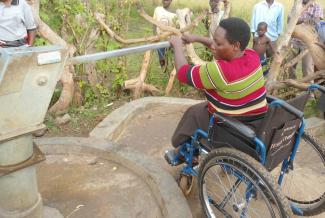Seorang perempuan pengguna kursi roda sedang mengoperasikan pompa tangan yang terletak di area pinggir beton