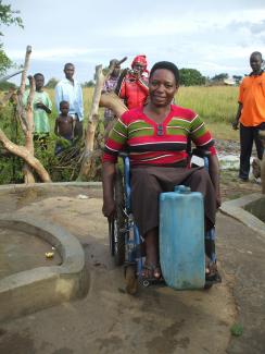 Une femme en fauteuil roulant à côté d'un puits, avec un bidon d'eau placé sur son repose-pied.
